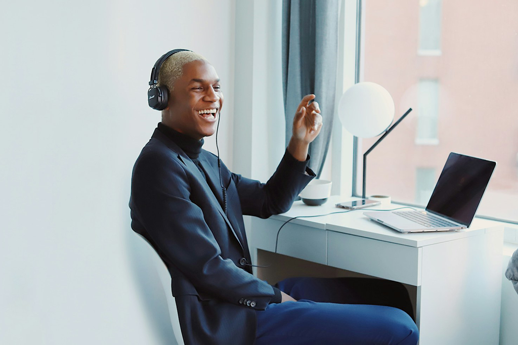 A black man in a suit sitting on an office chair in front of a window laughing with headphones in